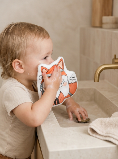 Bébé qui se nettoie la bouche de manière autonome près du lavabo avec un coton réutilisable en forme de renard. Accessoire de toilette bébé inspiré de la méthode Montessori.