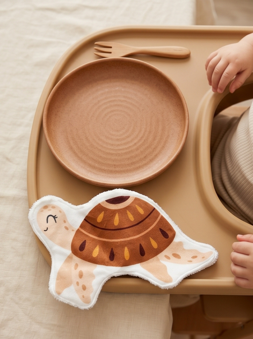 Grand coton lavable en forme de tortue marine posé sur le plateau d'une chaise haute pour enfant. Lingette lavable bébé artisanale et réutilisable pour débarbouiller après le repas.