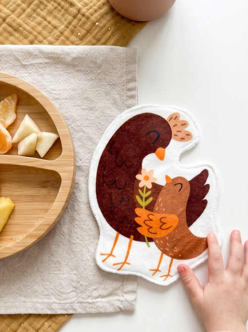 Grand coton lavable en forme de poule et son poussin posé à côté d'une assiette en bois compartimentée. Lingette lavable réutilisable artisanale pour débarbouiller les enfants après le repas.
