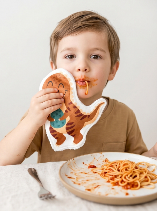 Débarbouillage autonome d'un enfant mangeant des spaghettis avec un coton réutilisable en forme de T-Rex. Accessoire de toilette ludique inspiré de la méthode Montessori.