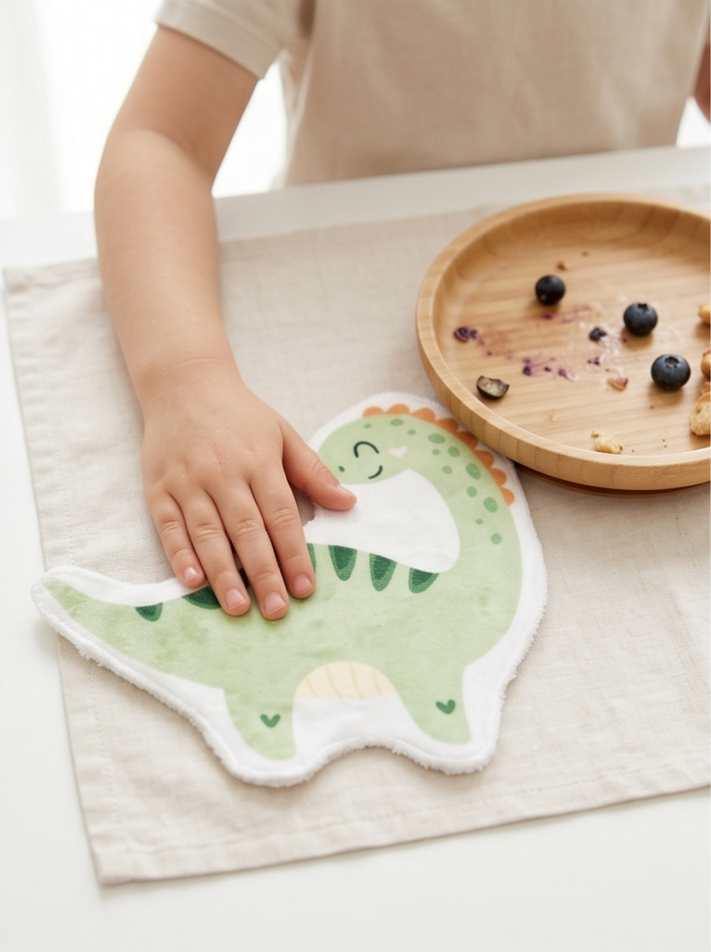 Main d'un enfant posée sur une lingette lavable en forme de dinosaure vert à côté d'un bol en bois. Coton lavable réutilisable écologique et zéro déchet pour enfant.