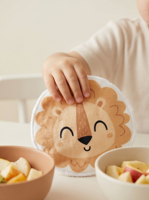 Enfant à table attrapant un coton réutilisable en forme de lion souriant à côté de bols de fruits. Accessoire de toilette ludique pour le débarbouillage autonome des petits.