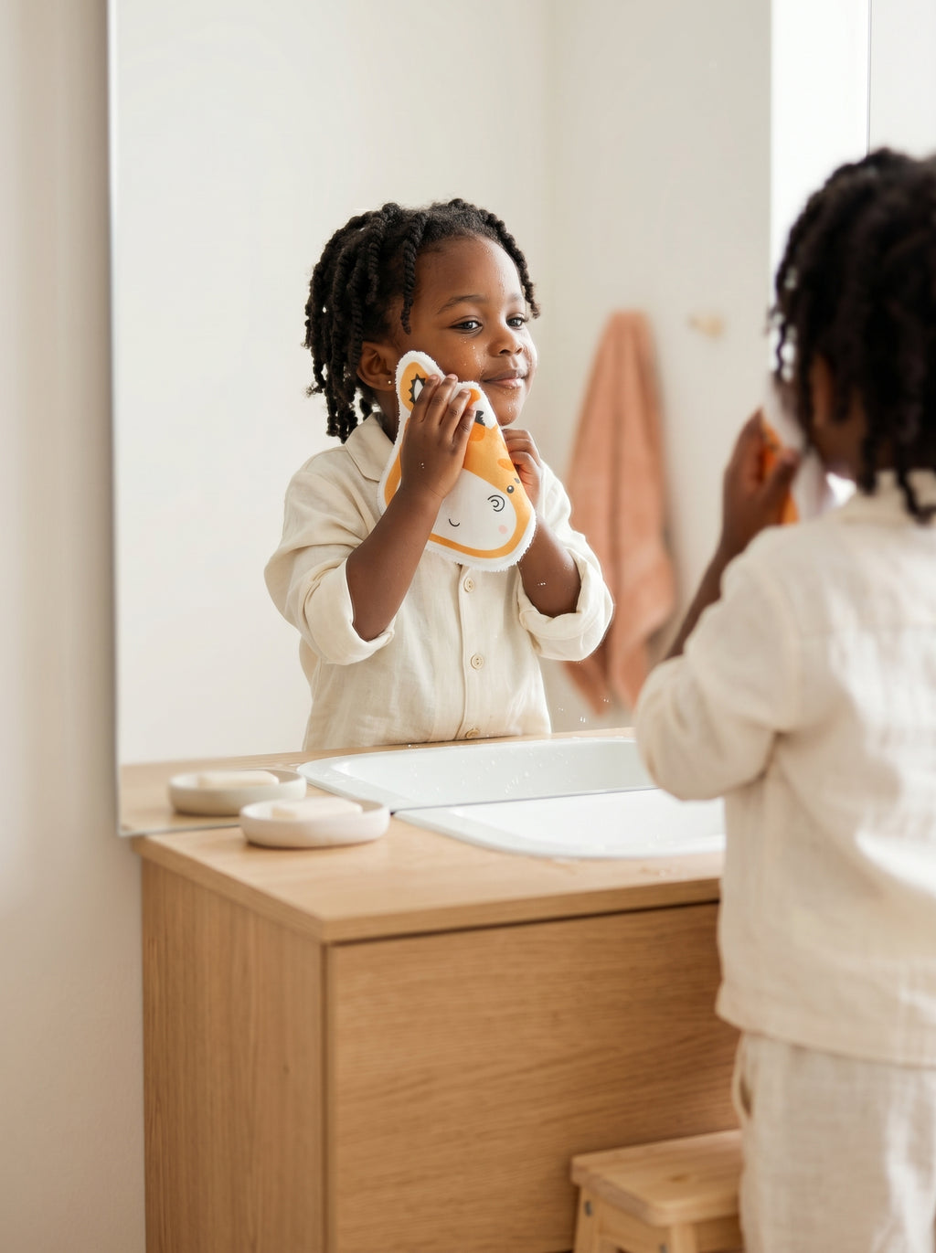 Enfant debout sur un marchepied dans une salle de bain style Japandi, utilisant un coton lavable réutilisable pour se débarbouiller en douceur. Alternative écologique zéro déchet.